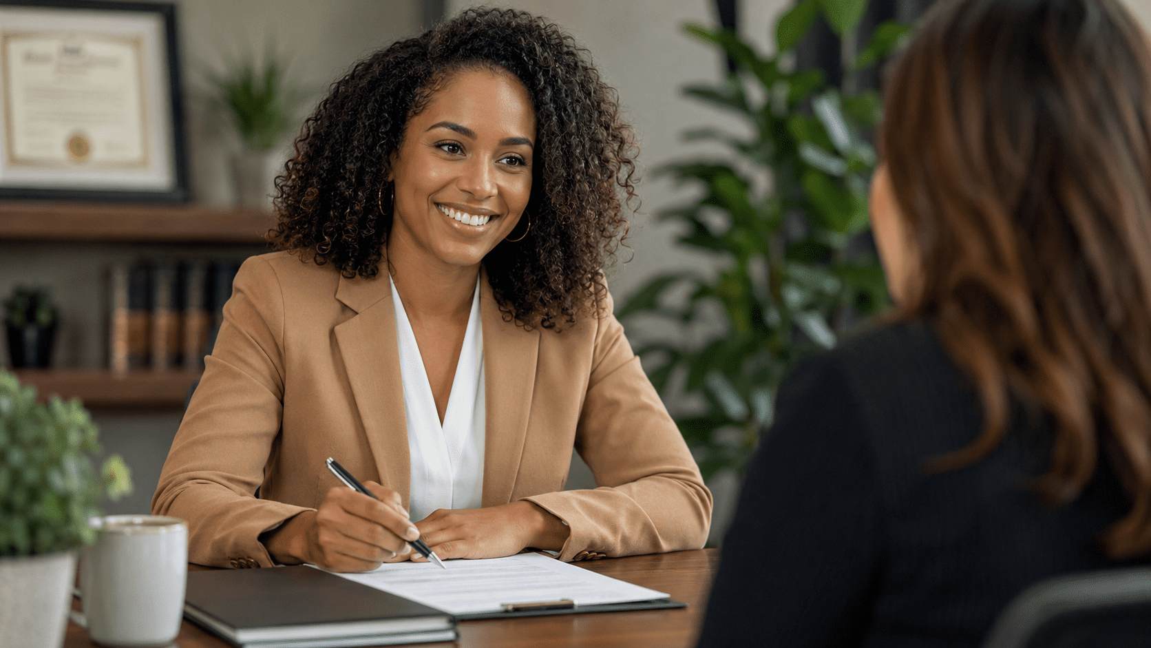 Two professional women in business attire collaborate at a modern reception desk, one holding a clipboard while the other speaks on a phone.